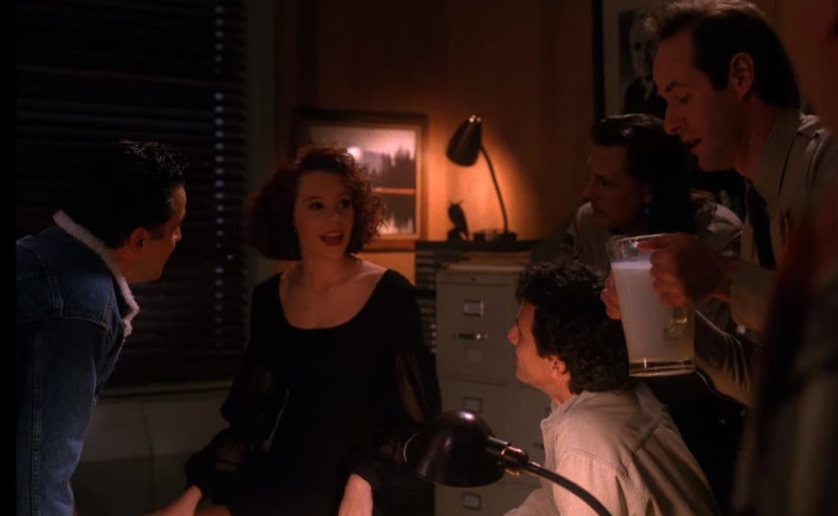 Lana Milford sits in the middle of a darkened office, with the only light behind her on a file cabinet, being surrounded and stared at by the men of the office
