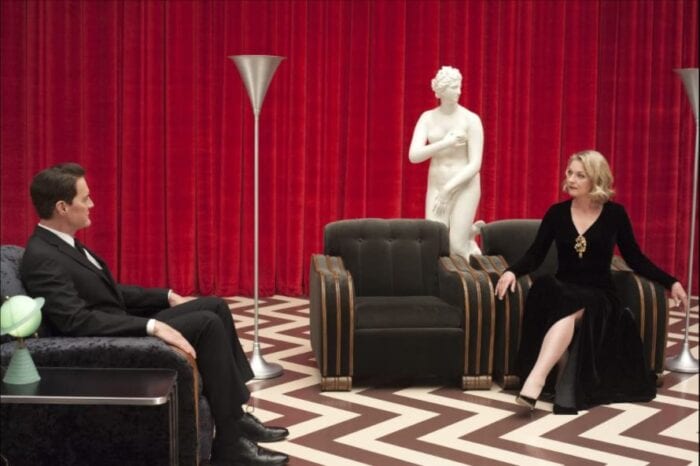 Dale Cooper (Kyle MacLachlan) sits across from Laura Palmer (Sheryl Lee) in the Red Room. Behind them is a statue, and there is a light in the shape of Saturn on the table.