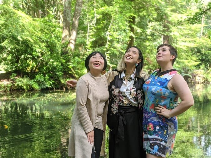 three smiling women stand in front of a pond and trees