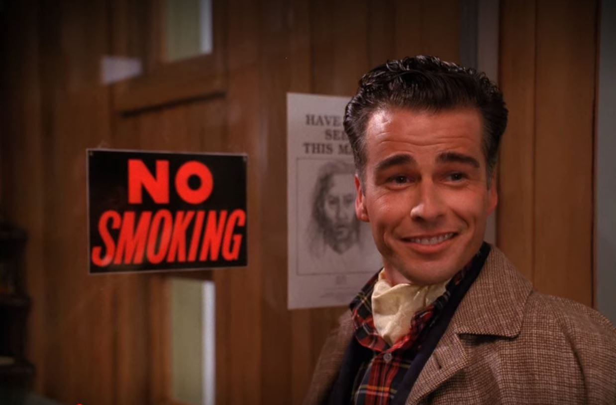Dick Tremayne smiles as he stands in front of a window in the Sheriff's station that has a No Smoking sign and a drawing of BOB posted on it