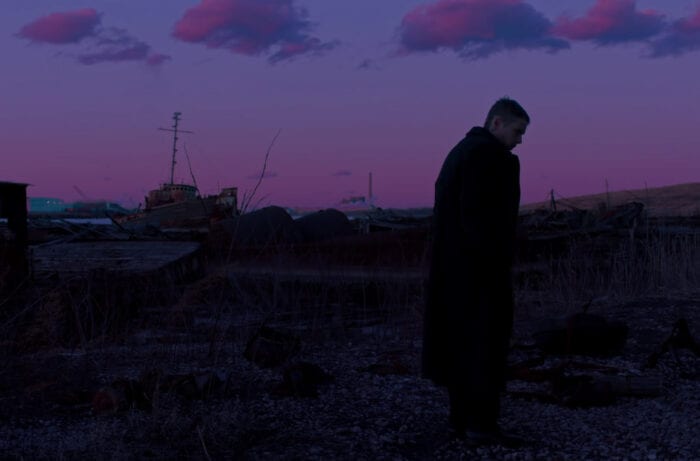 Reverend Toller stands outside in First Reformed. He is standing in the toxic waste site where Michael's ashes were scattered. The sky is a deep magenta. Toller is wearing a long black coat.