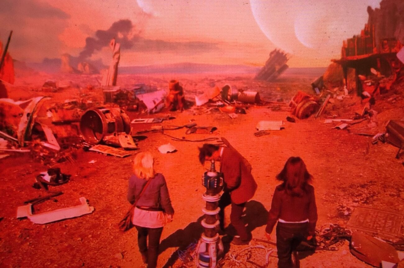 Jo, 11, and Sarah Jane stand around a mechanical device on a red-tinted alien planet