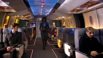 The hostess walks down the centre aisle of the train car in Doctor Who Midnight