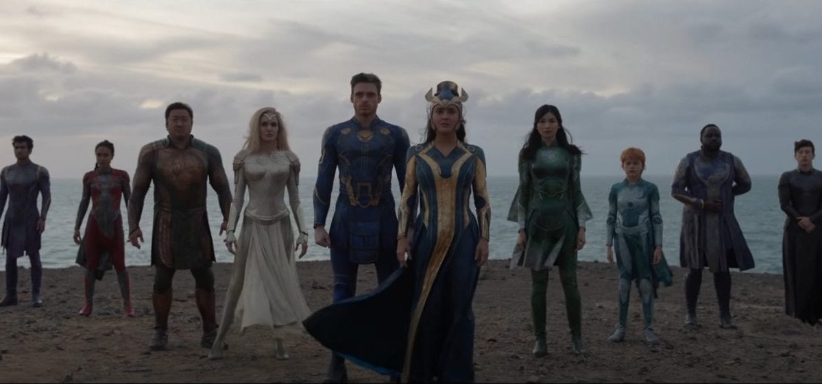 The Eternals stand in a V-formation on a cliffside with the ocean behind them
