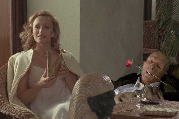 Almásy holds a rose in his mouth with a deformed face as Katharine sits next to him with a book in hand