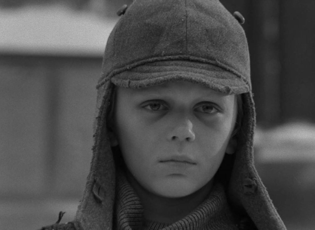 A young boy dressed in a Russian winter cap stares at the camera in this image from The Ascent.