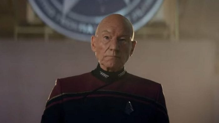 Jean-Luc Picard stands in front of a symbol on the wall behind him