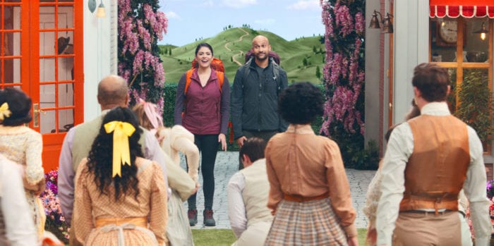 Mel (Cecily Strong) and Josh (Keegan-Michael Key) standing between two buildings facing the camera as the citizens of Schmigadoon stand looking back at them
