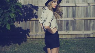 Taylor Swift stands in a yard with a wooden fence in the background, her hands on her hips as she wears shorts and a hat, looking back toward the camera