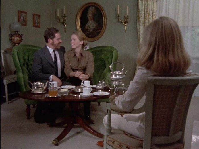 Johan and Marianne sit on a couch, close together, across a table holding tea from a woman in a chair
