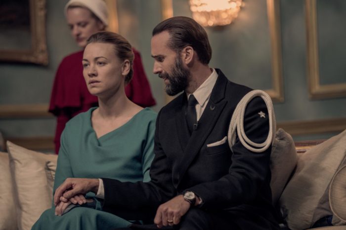 June, Serena, and Fred - The Handmaid's Tale. june is stood behind the waterford's, who are sat down