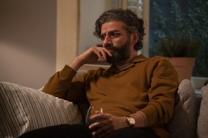 In this image from Scenes from a Marriage, Jonathan (Oscar Isaac) is depicted seated on a sofa stroking his beard and holding a glass of wine.