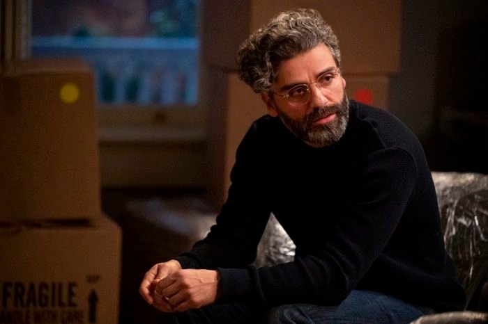 Jonathan (Oscar Isaac) is depicted wearing a black turtleneck and looking to his left, seated on a wrapped sofa.