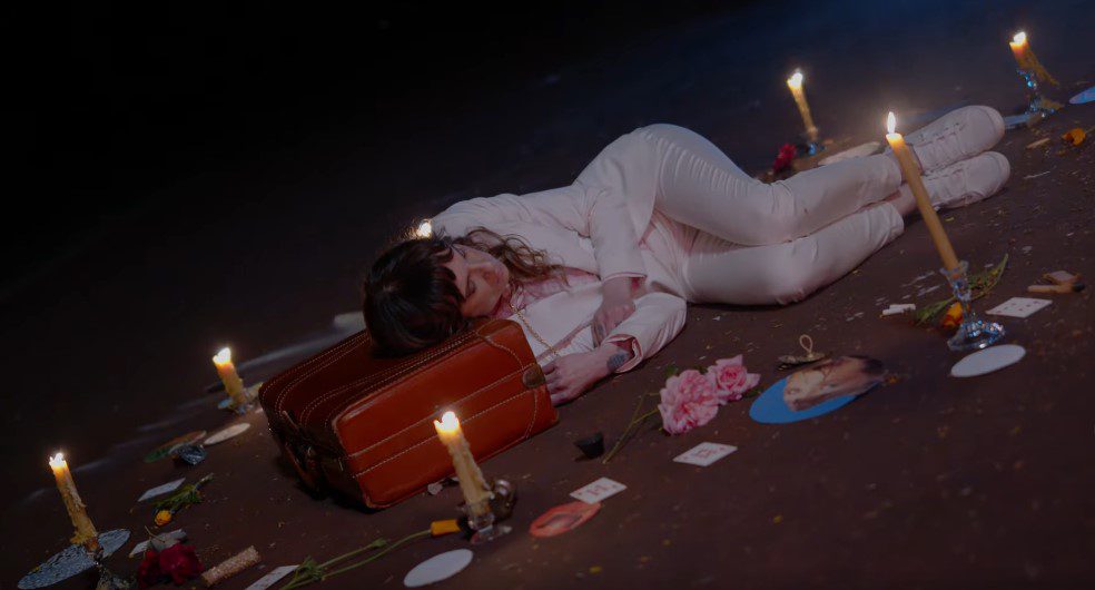 A woman wearing a white suit rests her head on a suitcase, surrounded by bright pieces of paper and lit candles (The woman is singer-songwriter Vonnie Kyle)