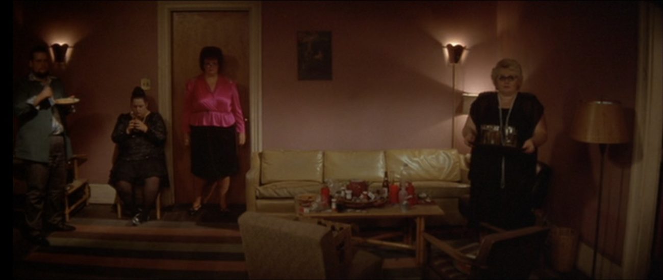 Three large women and one large man stand in a living room staring toward the camera; the man holds a plate of food and one woman holds a plate with several glasses of beer (from David Lynch's film Blue Velvet)