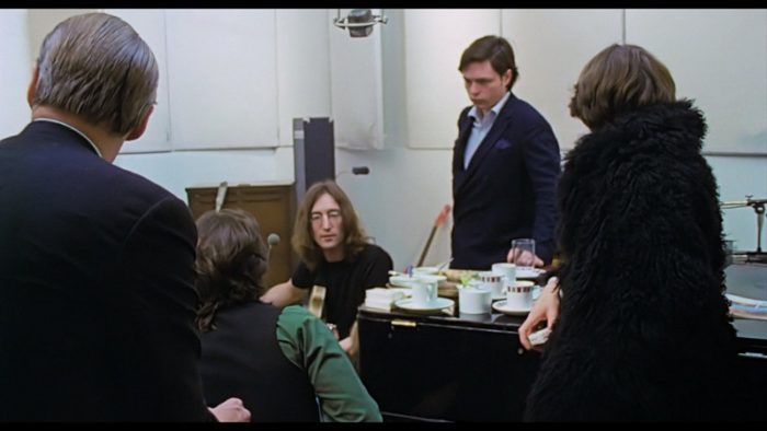 George Martin, Paul McCartney, John Lennon, Michael Lindsay-Hogg, and George Harrison sit near a piano