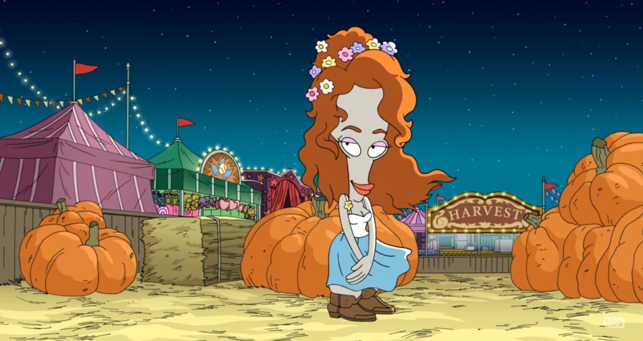 A bulbous-headed grey alien in an auburn wig wears a blue dress, brown boots, and flower crown; several large pumpkins and circus tents are in the background (Roger as Julia Rogerts in American Dad!)