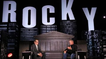 A host sits across from Sylvester Stallone under a large "ROCKY" stage set.