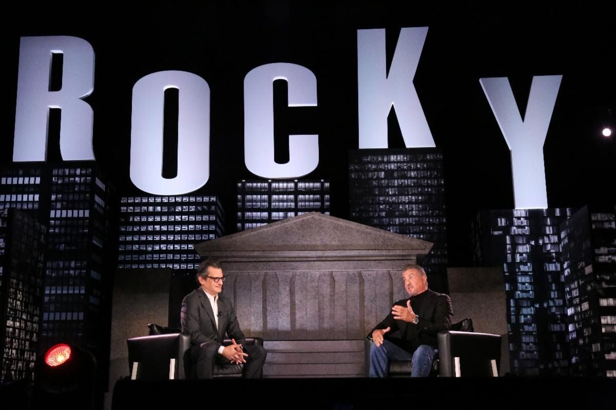 A host sits across from Sylvester Stallone under a large "ROCKY" stage set.