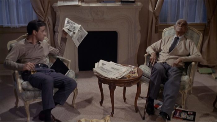 A young man and old man sit across from each other; the younger man is holding up a newspaper and the old man stares upward.
