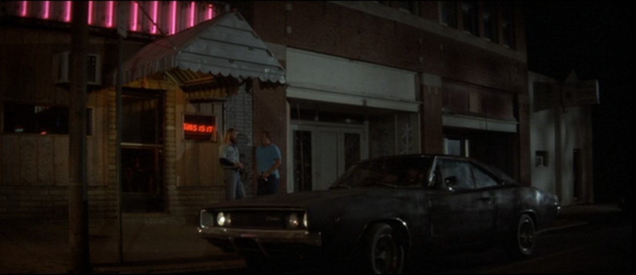 A black 1970s car parks in front of an old brick building. In the front window is a red neon sign that reads "This is it."