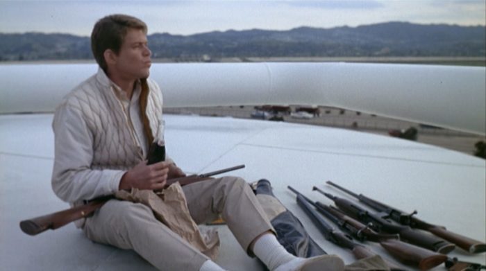 A young white man wearing khaki clothing sits atop a white oil tanker next to several rifles; he is holding a bottle of soda, looking off into the distance (Tim O'Kelly in Peter Bogdanovich's Targets).