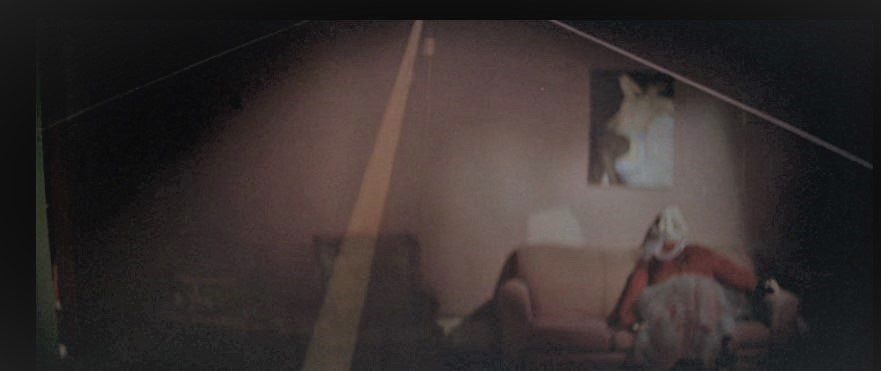 A woman wearing a red sweater, long skirt, and mask which covers her entire face and neck sits on a couch below a portrait of a naked woman; the yellow lines of a highway are superimposed onto the image (from David Lynch's film Blue Velvet)
