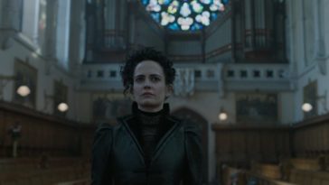 A woman with pale skin and black hair wearing all black walks through a cathedral; a large circle of stained glass is illuminated behind her (Eva Green in Penny Dreadful)