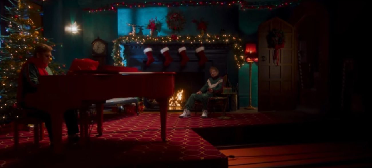 Elton John plays piano while Ed Sheeran sits by a mantleplace decorated with stockings