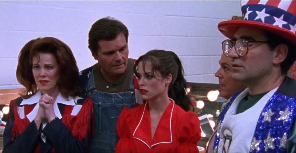 A woman with auburn hair, a man in overalls, a young woman in a bright red dress, and a man in glasses wearing a star spangled hat wear expressions of disbelief and sadness.