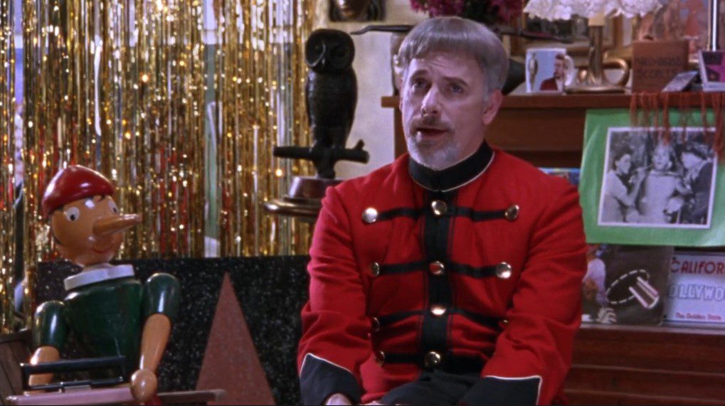 A man wearing a nutcracker outfit with a bowl haircut and goatee sits next to a large wooden puppet. 