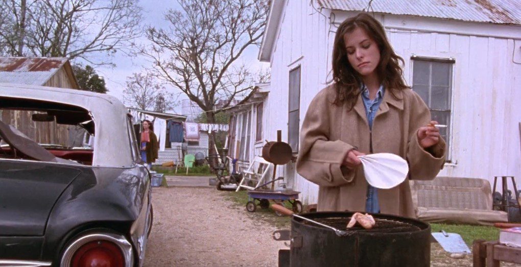A woman (Parker Posey) with brown hair wearing a beige jacket holds a cigarette in one hand and a fan in the other; she is cooking a single chicken wing on an outdoor grill.