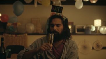 Jeevan (Himesh Patel) sitting in a wheelchair in a deparment store wearing a party hat and drinking champagne