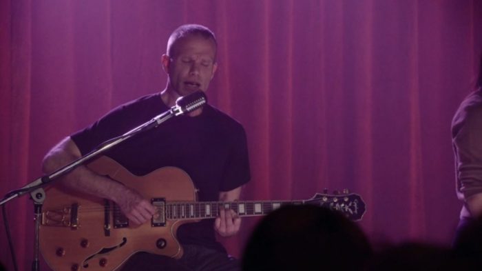 James Hurley strums his guitar and sings with his eyes closed, in front of a red curtain
