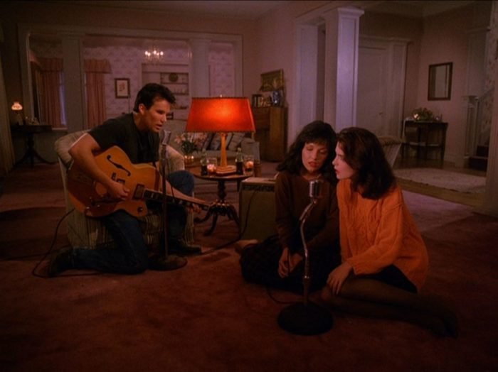 James kneels with a guitar in a living room as Maddy and Donna sit on the floor
