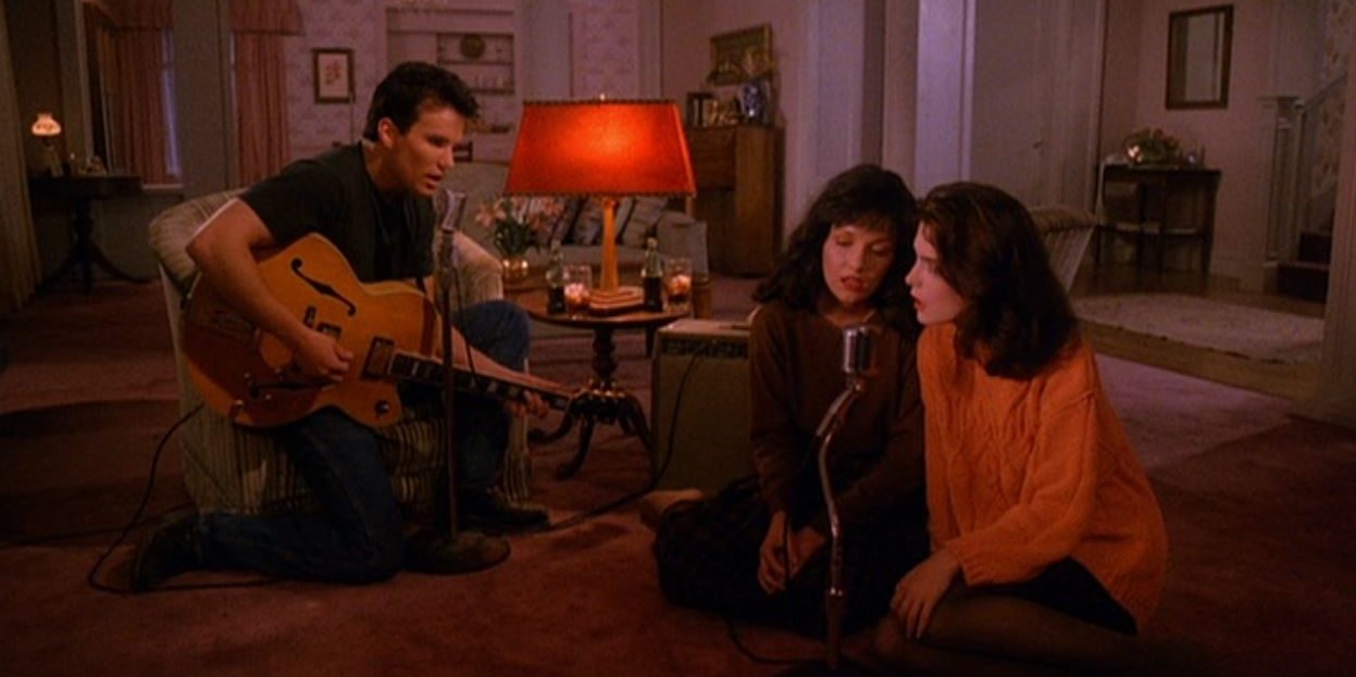 James kneels with a guitar in a living room as Maddy and Donna sit on the floor