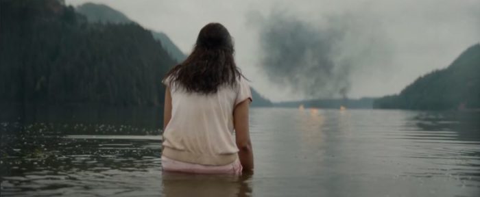 Lottie stands in the water of the lake, her back to us as she looks out at the smoke from the plane explosion