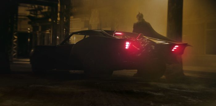 Batman stands next to a heavily modified car in the rain.