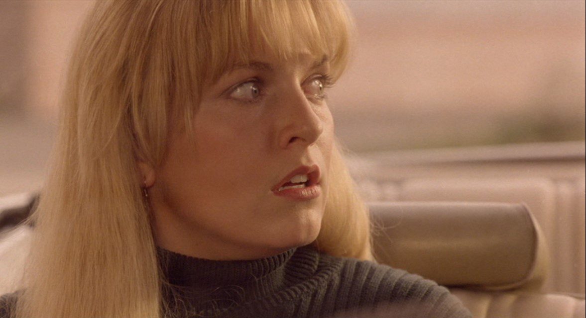 A close-up image of a young woman with long blonde hair in the passenger seat of a car. Her facial expression is one of shock.