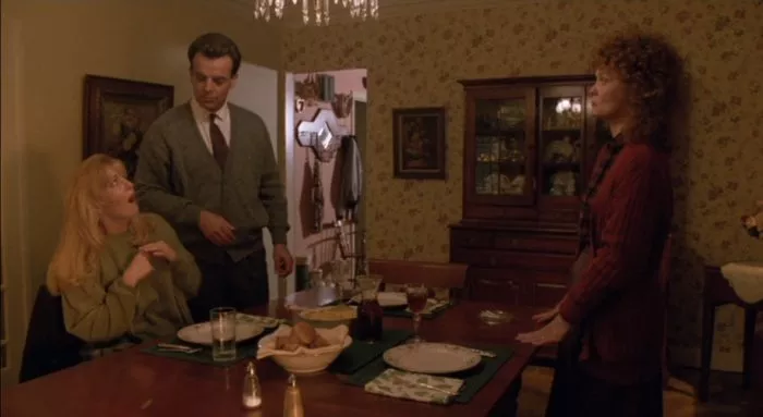 A man, Leland Palmer, and a woman, Sarah Palmer, stand across from each other at a large wooden table; a young woman, Laura Palmer, with long blonde hair sits at the table with her mouth hanging open in shock.