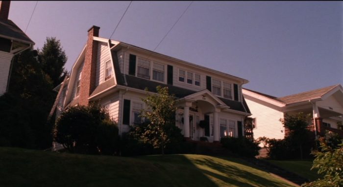 A large two story house sits atop a large green hill beneath a shadow. 
