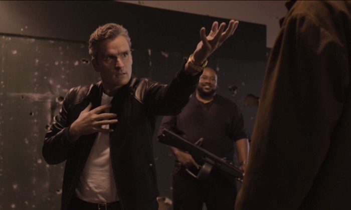 Balthazar Getty as Red, striking a pose and standing in front of a man with a gun in the background