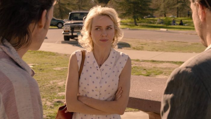 Naomi Watts as Janey-E Jones, standing facing two men in the foreground with her arms folded