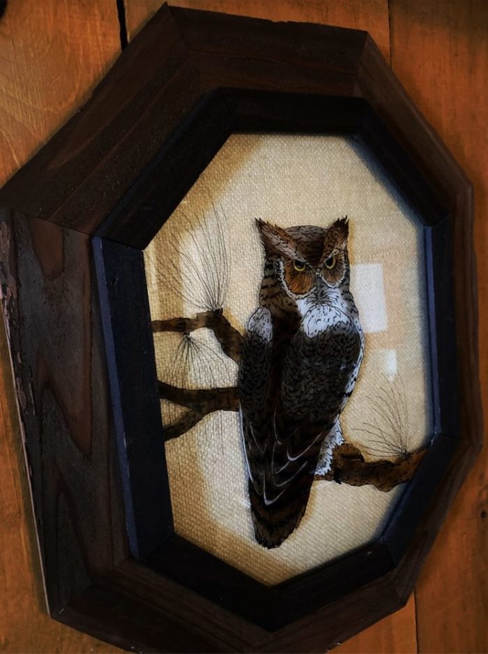 A cross stitched owl on a branch in an octagonal frame, on a wood paneled wall
