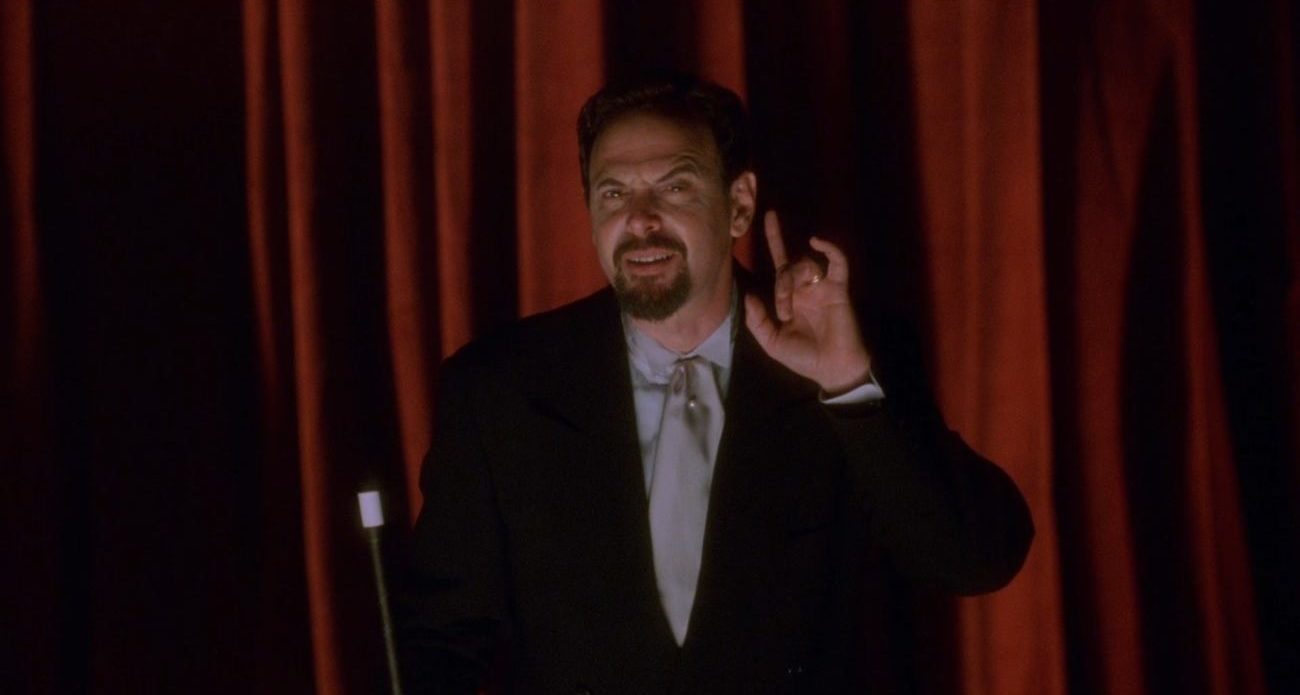 The Magician from Mulholland Drive, standing in front of red curtains and gesturing towards his ear to emphasize that we should listen