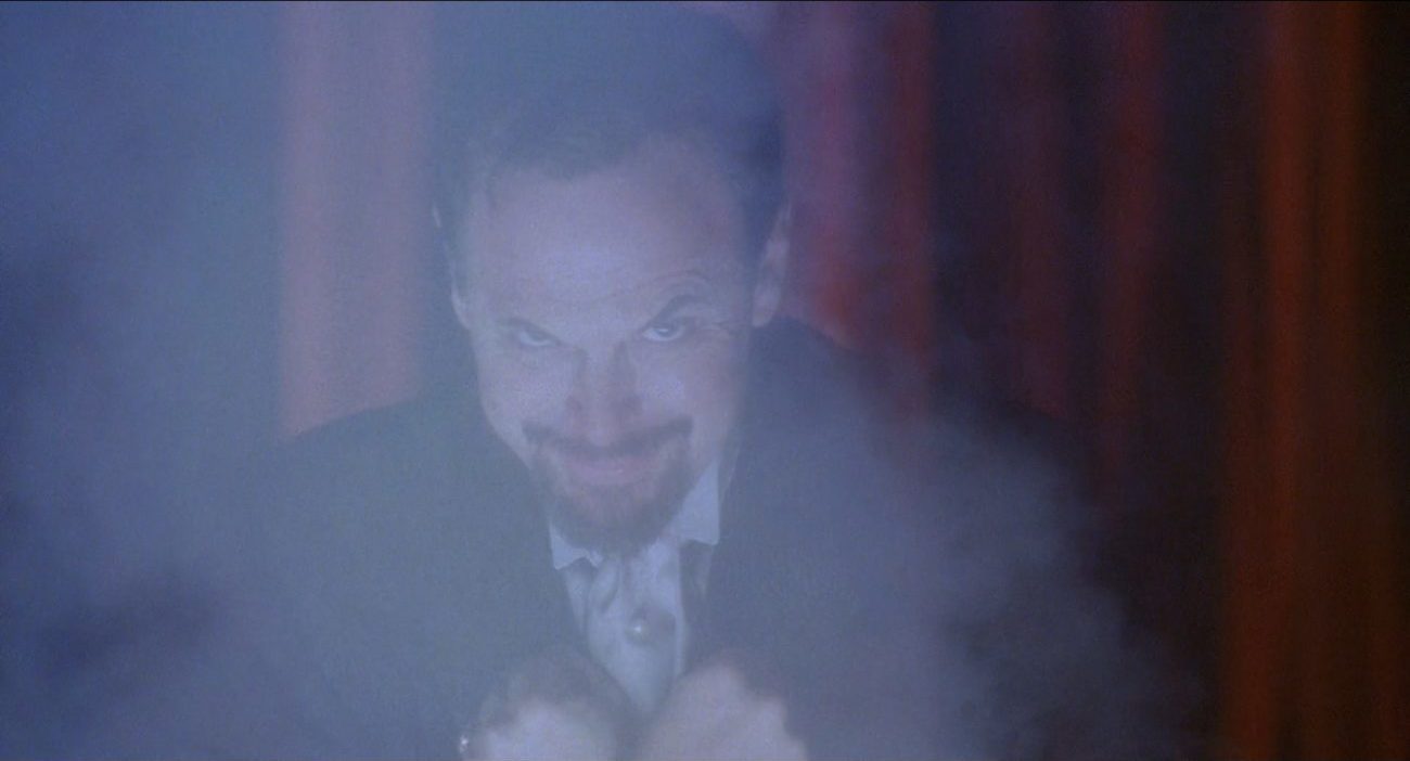 The Magician, staring at the camera with an unsettling smile on his face, his hands are crossed over his chest, smoke is partially covering his face and upper body, and there are red curtains behind him