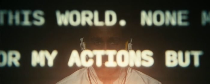 Milchick sits behind a projected screen bearing the words "this world. none" and on the second line "my actions but"