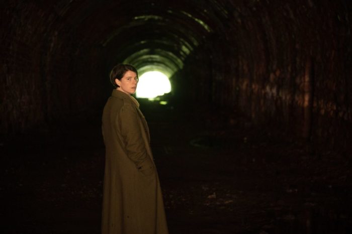 A person in a trenchcoat stands in a tunnel and looks back at the camera.