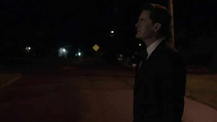 A dead end sign in the distance as Cooper looks at the Palmer house from the street.