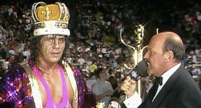 Bret Hart stand with an orange crown on and a sparkly jacket.
